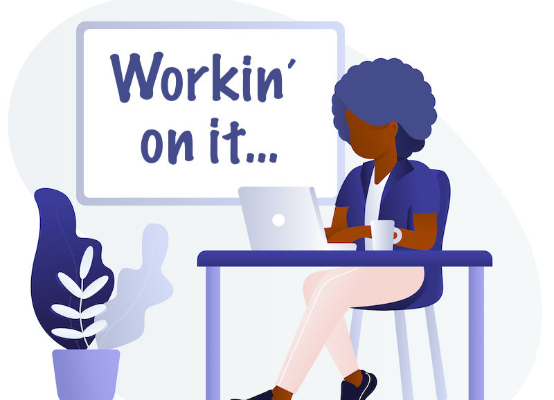 Woman working on a computer while sitting at a desk. "Workin' on it" is written on a white board behind her.